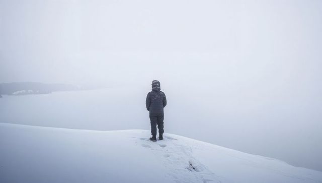 Standing figure on snowy ridge facing fog-filled expanse, footprints trailing into winter solitude