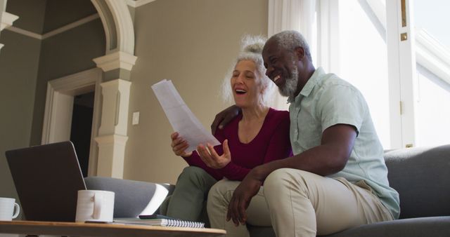 Senior couple sitting on sofa, smiling while reviewing documents together. They appear to be discussing important paperwork or enjoying a moment of shared accomplishment. Useful for themes around retirement, finance, joint decision-making, or family life.