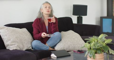 Mature woman relaxing on sofa sipping tea with notebook and smartphone, cozy home scene