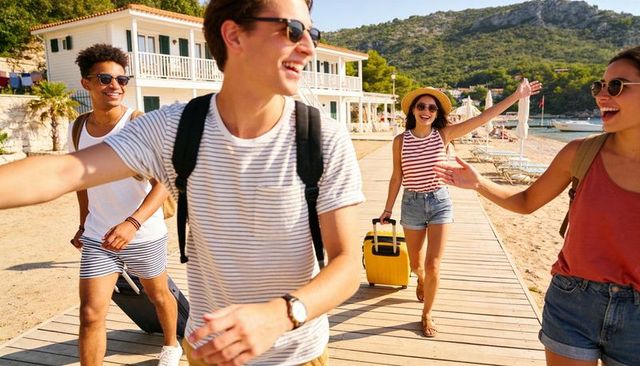 Young friends walking on seaside boardwalk pulling colorful suitcases, laughing and greeting