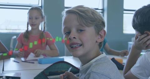 Smiling blond boy writing at classroom desk with stock chart overlay and classmates