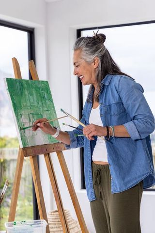 Senior woman painting green canvas on easel in sunlit studio, smiling and holding brushes