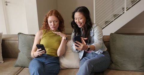 Two women sharing smartphone content on couch wearing smartwatches, smiling and laughing