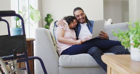 Interracial Couple Bonding on Sofa with Laptop, Wheelchair Visible