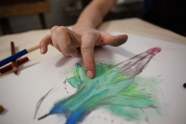 Close-up of a woman's hand shading a drawing with colored pencils on paper at a table. The drawing features green, blue, and red colors. Ideal for use in articles or advertisements about art, creativity, hobbies, and artistic techniques.