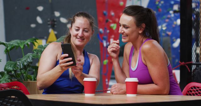 Two happy caucasian women laughing and looking at smartphone in cafe at indoor climbing wall. fitness and leisure time.