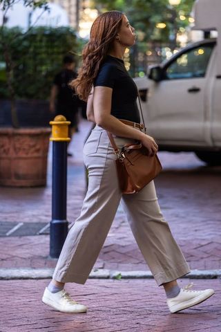 African american woman walking urban sidewalk wearing black top, white sneakers, brown bag