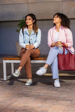 Two women sitting on bench holding coffee cups urban friends conversing in sunlight