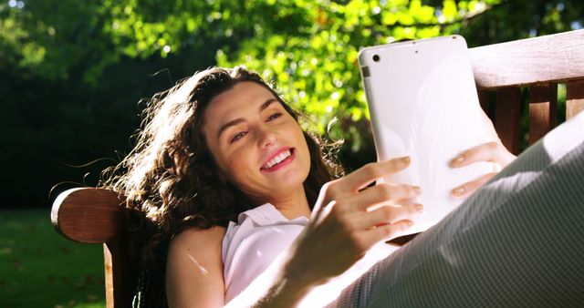 Smiling woman enjoying leisure time outside, using tablet on a bench in a sunny park. Great for concepts of relaxation, outdoor lifestyle, digital communication, and modern technology usage.