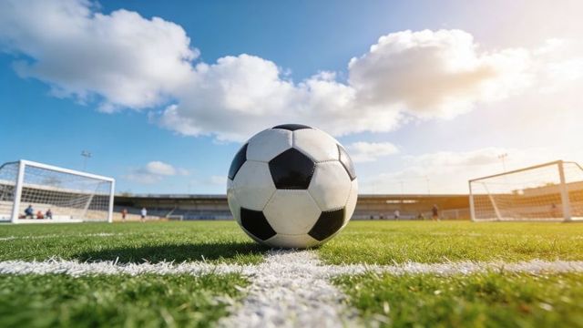 Low-angle soccer ball sitting on center line with stadium goals, players and sunlight
