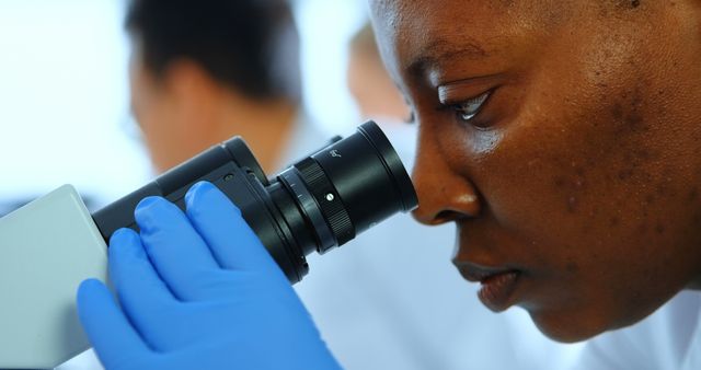 Scientist intently looking through microscope, wearing blue gloves, blurred background. Ideal for use in educational materials, scientific publications, research presentations, healthcare promotions, and articles on laboratory work.