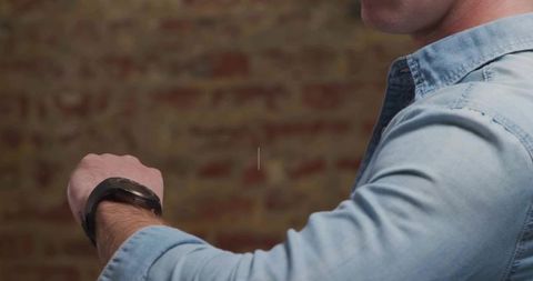 Man wearing denim shirt presenting round-faced smartwatch on wrist against brick wall