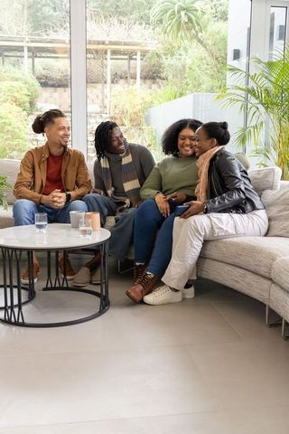 Diverse friends laughing on sectional sofa sharing smartphone and coffee in living room