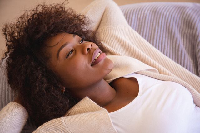 African American woman lying on a couch with a relaxed smile, eyes closed, enjoying leisure time at home. Ideal for use in lifestyle blogs, self-care articles, home decor advertisements, and mental wellness promotions.