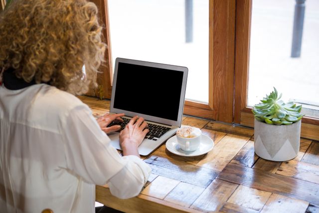 Young woman with curly hair typing on laptop in a cozy coffee shop. She is seated at a wooden table with a cappuccino and a potted plant nearby. Ideal for illustrating remote work, freelancing, modern work environments, and casual work settings.