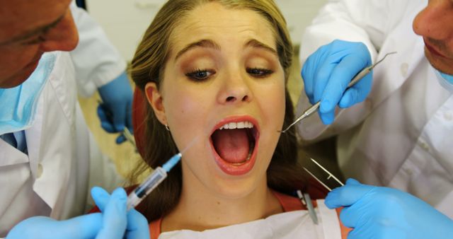 A young Caucasian woman is receiving dental treatment from professionals, a dentist and an assistant, with copy space. She appears anxious as the dentist prepares to administer an injection, highlighting the common fear of dental procedures.