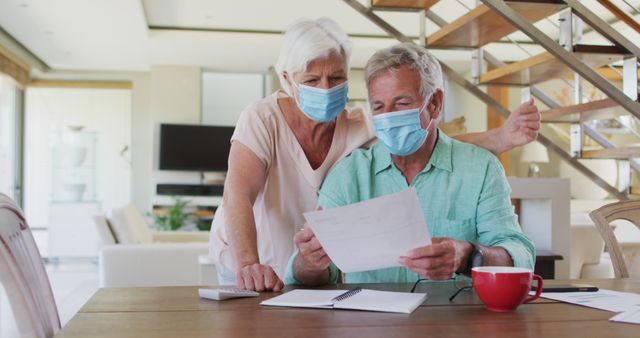 Senior couple wearing masks while reviewing documents at home. This can be used to depict themes of health safety during the pandemic, financial planning, home lifestyle, and elderly care. Ideal for articles or promotional materials related to COVID-19 safety measures, retirement planning, elder care, and family health precautions.