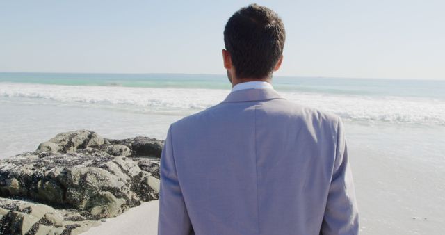 Businessman standing on the beach looking at the ocean present an image of contemplation, possibly reflecting on work or life. The scene can be used to illustrate themes of work-life balance, relaxation, corporate retreat, or future planning. Ideal for articles, advertisements, or corporate materials focusing on professional life and mental well-being.