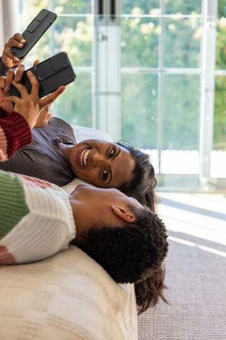 Multicultural couple lying on bed using smartphones, laughing together in sunlit cozy home