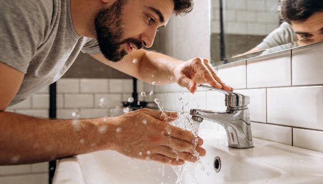 Bearded man washing hands at bathroom sink with splashing water and mirror reflection