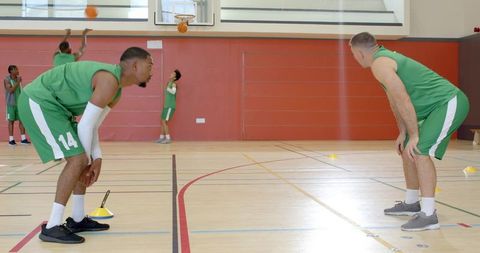 Diverse basketball team practicing drills in gymnasium, shooting, sprinting, coaching