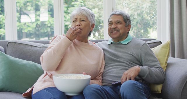 Senior couple sitting on sofa and enjoying a movie, sharing a bowl of popcorn. This warm and comforting scene can be used in advertisements, blogs, or articles focused on senior living, retirement, family leisure time, and home comfort.