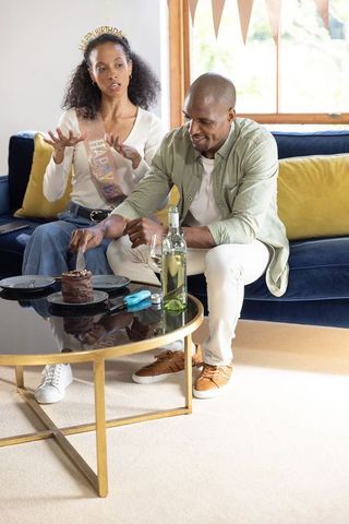 Diverse couple celebrating birthday at home cutting chocolate cake on navy velvet sofa