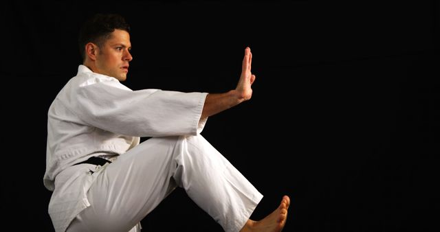 Man wearing martial arts gi practicing high kick and hand motion in studio with black background. Suitable for promotion of martial arts training, fitness, self-defense classes, discipline and focus advertising.