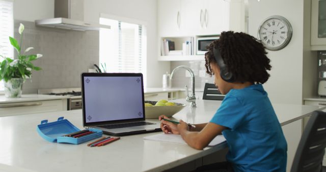 Young boy is sitting at kitchen island focusing on learning through his laptop. He is wearing headphones, with school supplies spread around, signifying dedicated study time. Ideal for use in educational technology, homeschooling resources, remote learning platforms, back-to-school promotions, child development materials, and e-learning content. Showcases intersection of modern home spaces with contemporary education trends.