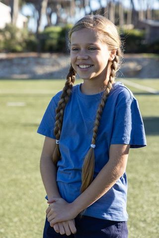 Smiling schoolgirl with long braids on sunlit sports field wearing blue t-shirt