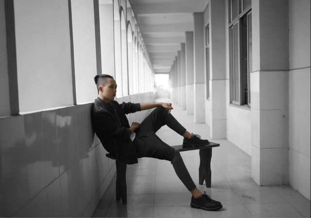 Young man relaxing on bench in corridor in modern building, dressed in black outfit. Perfect for themes related to fashion, modern lifestyle, architecture and solitary moments.