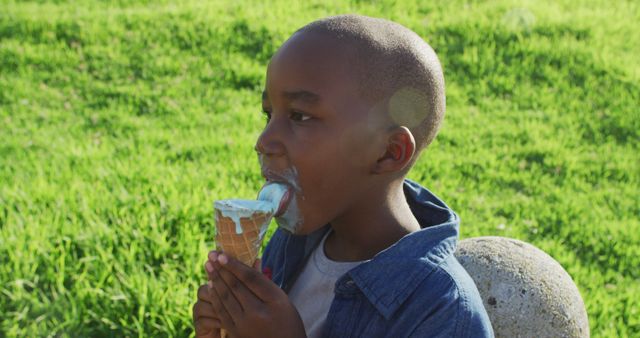 Boy licking ice cream cone with grin on his face, sitting in grassy field during bright sunny day. Ideal for ads and articles on childhood, summer activities, and sweets.