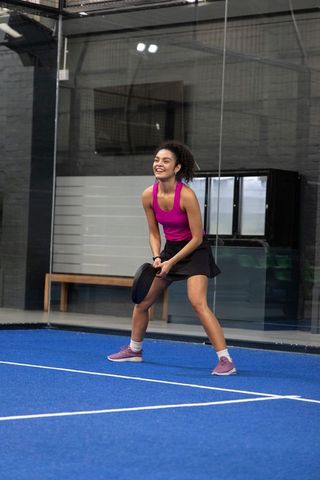 Smiling african american woman playing padel on indoor blue court wearing magenta top