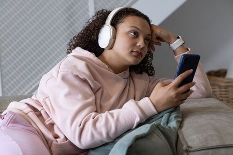 African american woman relaxing on sofa with headphones and smartphone in pink loungewear