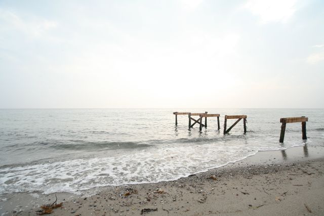 Waves gently lap the shore near a dilapidated wooden pier extending into the calm sea. The sky is overcast, adding to the serene atmosphere. Perfect for conveying tranquility, solitude, or nature themes. Ideal for beach holiday promotions, mindfulness and relaxation content, or seaside retreats marketing.