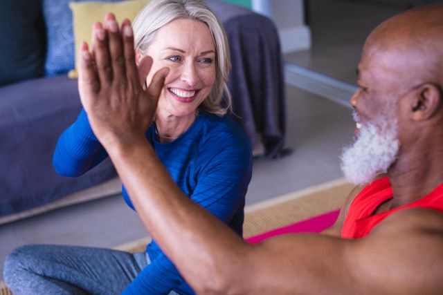 Happy senior diverse couple in exercise clothes practicing yoga together, highfiving. healthy, active retirement lifestyle at home.