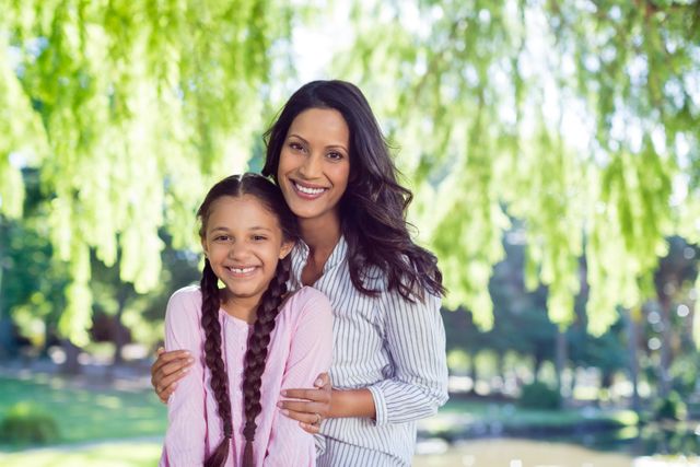 Mother and daughter smiling together in a park with lush greenery in the background. Ideal for use in family-oriented advertisements, parenting blogs, and promotional materials for outdoor activities or family events.
