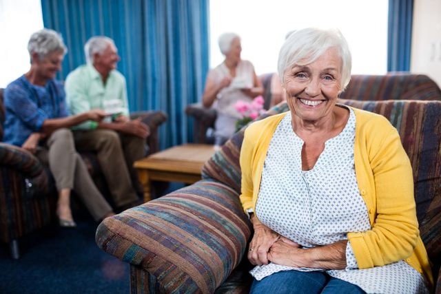 Senior woman smiling while sitting on a couch in a retirement home. In the background, other elderly individuals are socializing and enjoying each other's company. This image can be used for promoting senior living communities, elderly care services, and retirement planning.