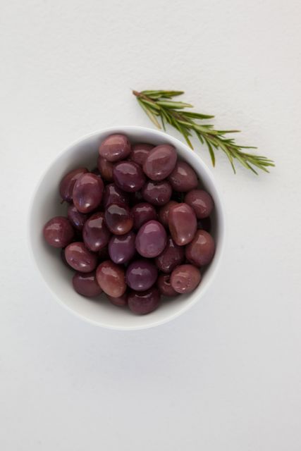 Overhead view of brown olives in bowl by herb on white table