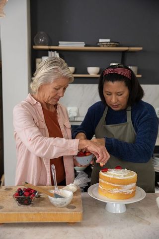 Diverse mature friends decorating sponge cake with strawberries in cozy modern kitchen
