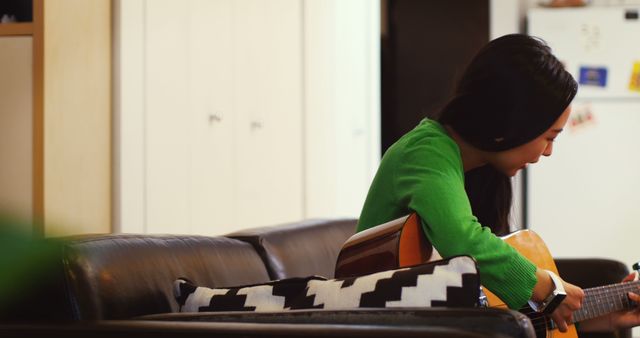 This image of a woman practicing guitar while sitting on a couch in a cozy living room can be used for promoting music lessons, lifestyle blogs, or advertisements related to home furnishings. It captures a serene and personal moment, highlighting themes of relaxation, creativity, and indoor leisure activities.