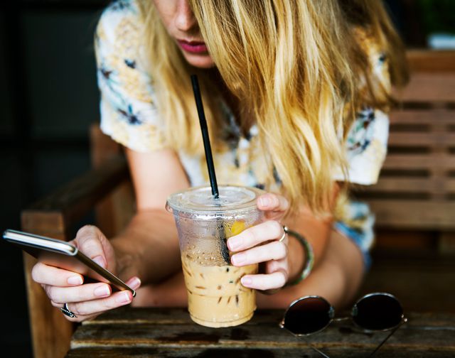 Young woman with blond hair sitting on wooden bench, holding iced coffee with one hand, texting on smartphone with other hand. Perfect for illustrating modern technology use, lifestyle blogs, summer activities, and coffee shop atmospheres.