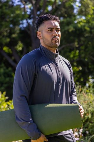 African american man holding rolled yoga mat in sunlit park wearing dark activewear