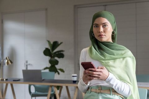 Young professional muslim woman wearing green hijab holding smartphone in modern office