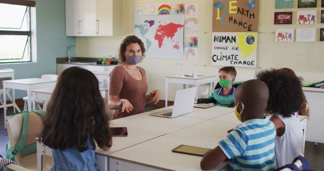 Teacher educating diverse group of children in a classroom, discussing climate change. All wearing masks and following Covid-19 protocols. Ideal for concepts related to education, diversity, climate awareness, Covid-19 adaptations in schools, and learning environments.