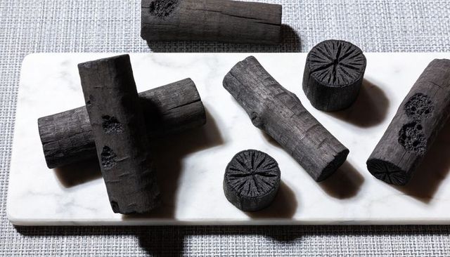 Charcoal logs on marble slab casting bold shadows, showing textured briquettes and wood rings