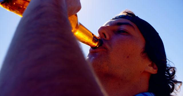 Young adult drinking beer outdoors on a bright sunny day. Close-up angle captures the concept of leisure and enjoyment in a casual setting. This image is suitable for use in advertisements for alcoholic beverages, blog posts about outdoor activities, summertime relaxation themes, or promotional materials for casual gatherings.