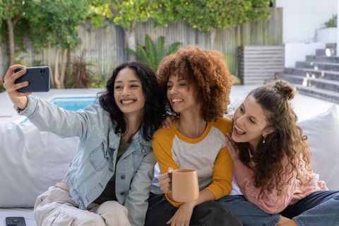 Diverse women taking selfie by pool laughing on outdoor sofa holding coffee mug