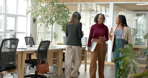 Diverse women walking and talking in modern open-plan office holding laptop and portfolio