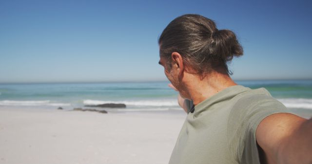 Middle-aged man with long hair tied in a ponytail relaxing on a sandy beach on a sunny day. He is pointing towards the horizon with the ocean waves in the background. Ideal for promoting travel destinations, beach vacations, leisure activities, and holiday packages.